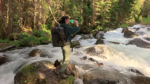 Female Traveler with a Backpack Drinking Water in Nature in the Forest Near a Mountain River
