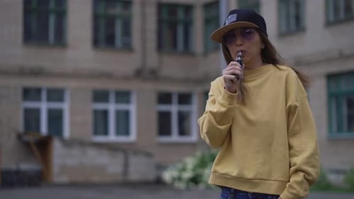 Bonita joven hipster con gorra negra y sudadera con capucha amarilla Vape Ecig. Dispositivo de eccig popular de Girl Vape