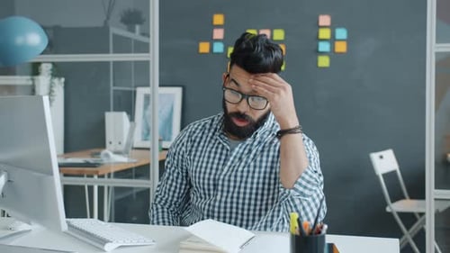 Middle Eastern Man Working in Office Using Computer and Reading Notes Feeling Headache