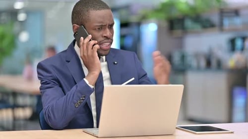 African Businessman with Laptop Angry on Smartphone in Office