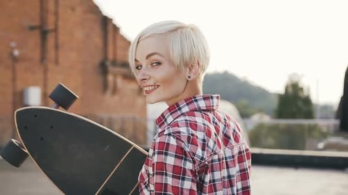 Beautiful Blonde Girl Walking with Long Skateboard on the Modern Building Roof