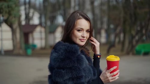Beautiful woman with a plastic cup of coffee in the park. Portrait of brunette girl in fur coat posi