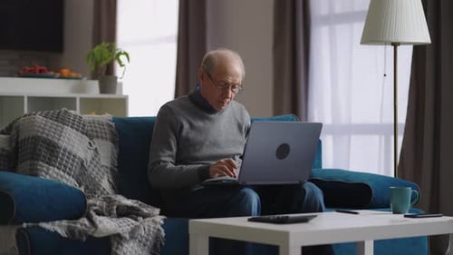 Senior Man Uses Laptop Sitting on Couch