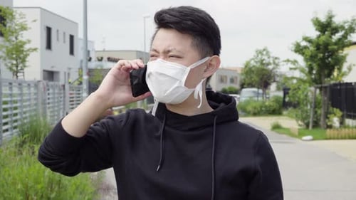 A Young Asian Man in a Face Mask Talks on a Smartphone in an Empty Suburban Area