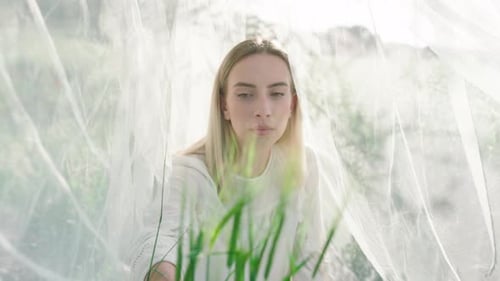 Thoughtful young woman surrounded by mosquito net sitting on farm field during sunset