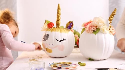 Little girl and her mother painting face of a unicorns on her craft pumpkin for Halloween.
