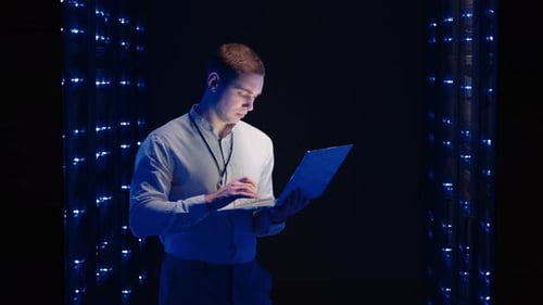 IT Specialist Works with a Laptop in the Server Room Looking at the Cabinets with Servers