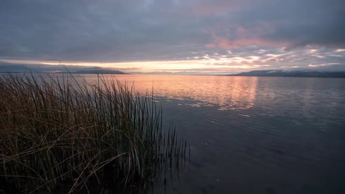 Time lapse of sunset over glassy lake looking past reeds