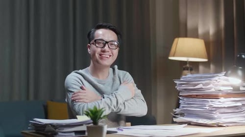 Asian Man Crossing Arms And Smiling To Camera While Working With Documents At Home