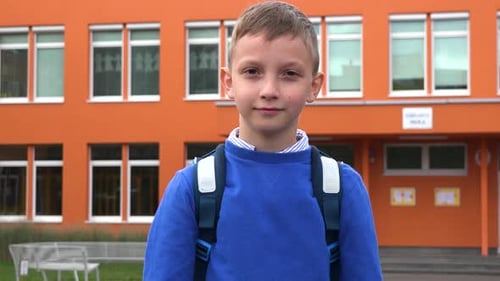 A Young Boy Smiles and Waves at the Camera - an Elementary School in the Background