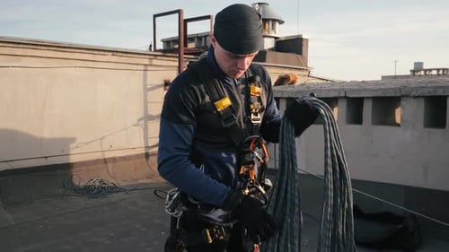 Alpinista industrial em uniforme fica no telhado de um prédio alto e pega uma longa corda de segurança