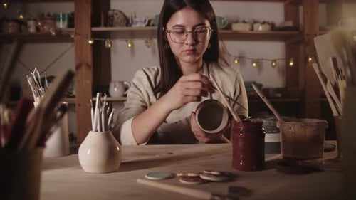 Young Woman Potter Drawing a Design on the Ceramic Mug with a Brush Then Looks in the Camera