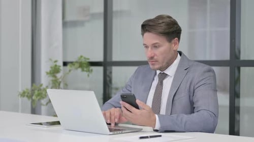 Middle Aged Man using Smartphone while using Laptop in Office