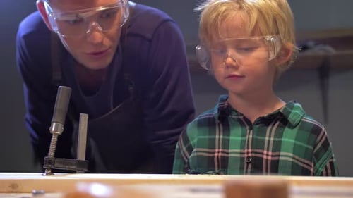 Boy Blows Away Shavings From a Wooden Board in a Workshop