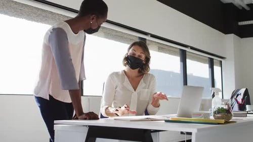 Two diverse female colleagues wearing face masks looking at laptop and discussing in office