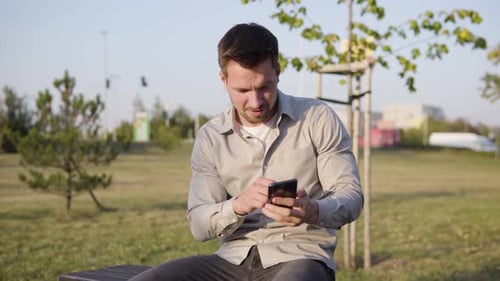 A Young Caucasian Man Takes Selfies with a Smartphone As He Sits in a Park in an Urban Area