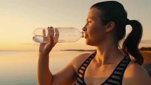 Woman Drinks Water by Ocean at Sunset