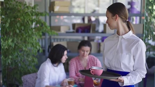 Junge schlanke Kellnerin mit Kaffeetassen auf Tablett blickt zurück auf Kunden, die am Tisch sitzen