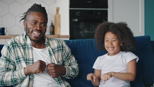 Happy Playful African American Father and Daughter Dancing Together and Laughing Sitting on Sofa at