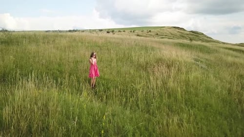 Yong Woman with Long Hair in Red Dress Walking in Summer Field with Tall Green Grass on Rural Hill