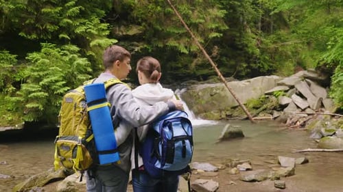 Crane Shot A Couple of Tourists with Backpacks Admire the Beautiful Waterfall and the Mountain River