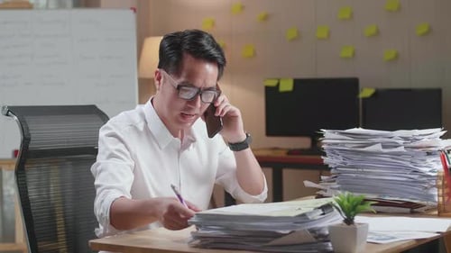 Asian Man Talking On Phone And Taking Note While Working Hard With Documents At Office