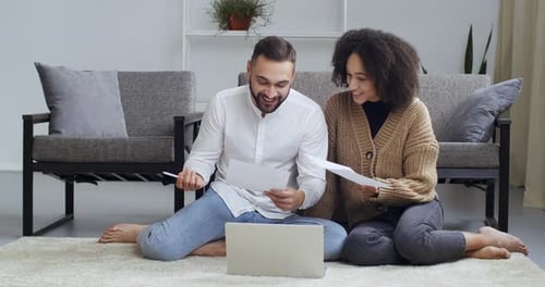 Multiethnic Couple Sitting at Home Living Room Floor in Front of Laptop Holding Papers in Hands