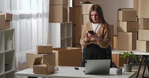 Woman Using Smartphone at Desk Surrounded by Boxes