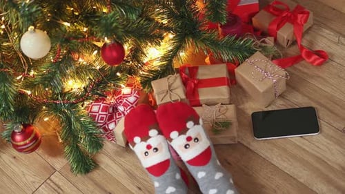 Feet in Funny Christmas Socks Santa Face Wooden Floor Under Fir Tree Gift Boxes