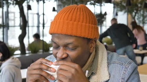 Black Guy in Jacket and Hat Tastes Delicious Burger in Cafe