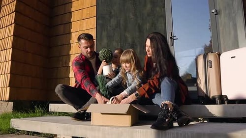 Happy Joyful Family Sitting on Steps and Getting from the Box Green Plants on the Moving Day