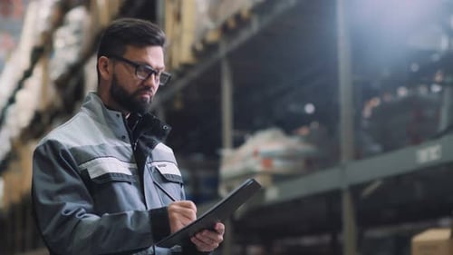 Man in Gray Uniform with a Folder Counts the Boxes in the Warehouse