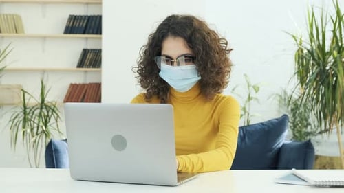 Young Business Woman in a Medical Mask Works From Home at the Computer During Selfisolation and