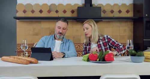 Hombre mostrando algo en una tableta a su hermosa esposa rubia que prepara una cena familiar