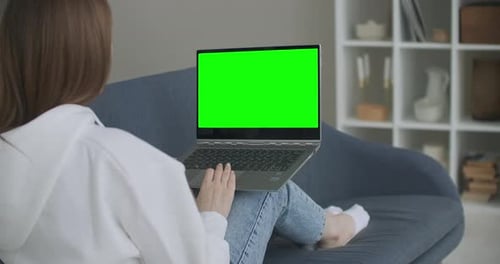 Woman at Home Sitting on a Couch Works on a Laptop Computer with Green Mock-up Screen. Coronavirus