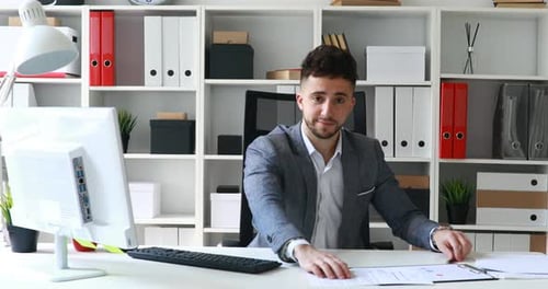Geschäftsmann in grauer Jacke sitzt am Tisch im weißen Büro und bietet Dokumente an