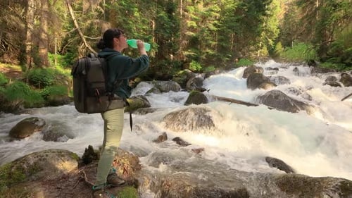 Female Traveler with a Backpack Drinking Water in Nature in the Forest Near a Mountain River