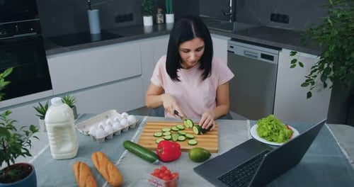 Morena en la cocina moderna y cortando pepino para ensalada eligiendo simultáneamente el programa en la computadora portátil