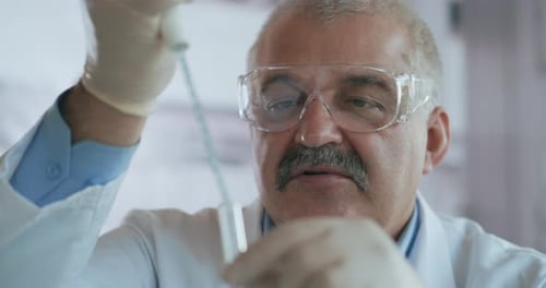Rack Focused Shot of Male Caucasian Scientist in Medical Gown and Goggles Pouring Red Liquid Into