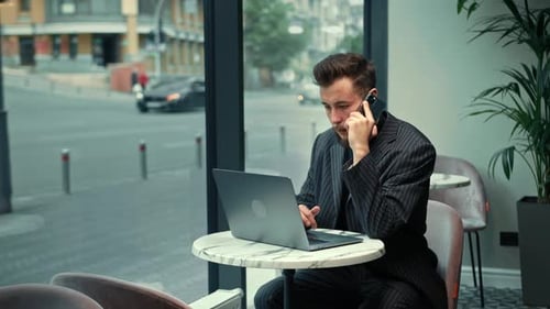 Bearded man is working on laptop speaks by the phone in a coffee shop. Freelancer, businessman.