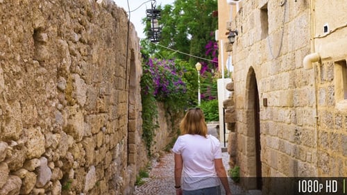 Following young woman walk down narrow cobblestone street with stone walls and houses