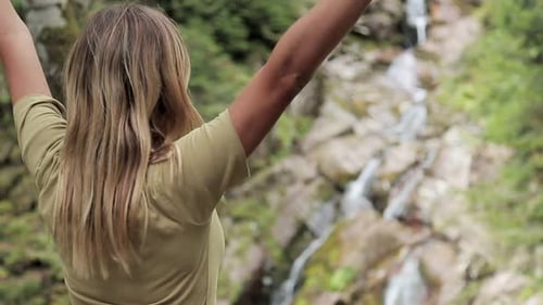 Rear View of Young Woman Standing in Front of Waterfall with Her Hands Raised. Female Tourist with