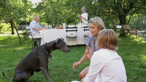 Children in garden playing with dog, parents at table in the background eating