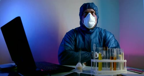 un hombre con ropa protectora azul con capucha, guantes y respirador está sentado en una mesa con