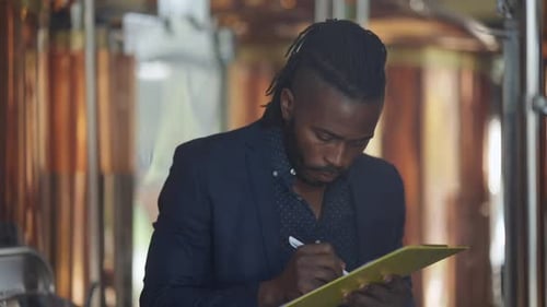Un brasseur afro-américain expert vérifie la qualité de la bière, remplit de la paperasse debout dans de grands réservoirs