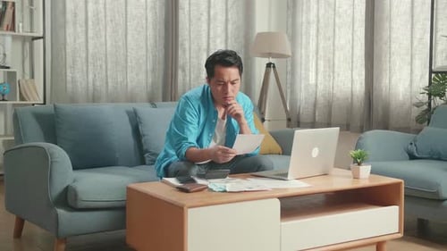 Asian Man Looking At The Bill And Thinking Before Typing On A Laptop At Home