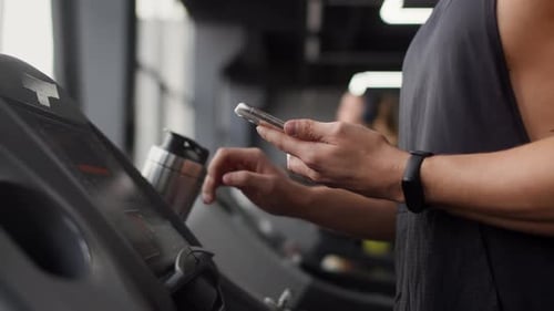 White Man in the Gym Turns on the Treadmill and Walks Along It Holding Smartphone in His Hand