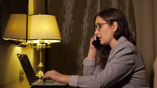 Woman in a Business Suit in the Evening Works at a Laptop and Talks on the Phone