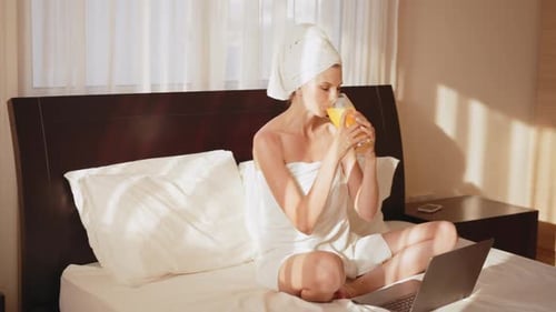 Blonde Woman Drinks Juice on Bed with Laptop