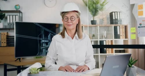 Arquitecta con casco sentada en el lugar de trabajo frente a la cámara en una oficina de diseño moderno